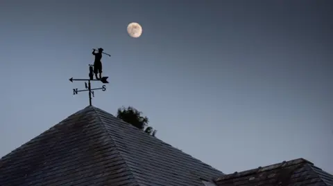 Alan Rankin A weather vane on the roof of a house has a silhouette of a golfer hitting a shot. Just above the figure is the moon. The sky is dark and starless.