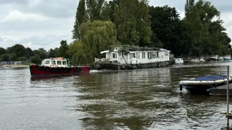 Elmbridge Borough Council A white boat is being towed away by a red boat. 