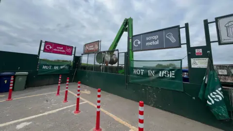 The Hempsted Household Recycling Centre. Bays for 'small electricals', 'wood and timber', 'scrap metals', and 'general waste'. Orange and white striped bollards sit in front of them.