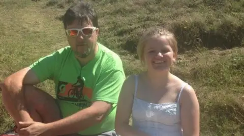 Eve Thomas Eve, a young teenage girl wearing a white dress and a ponytail, sits on a grassy hill in the sunshine with her father Nigel, who is wearing a green t shirt and sunglasses