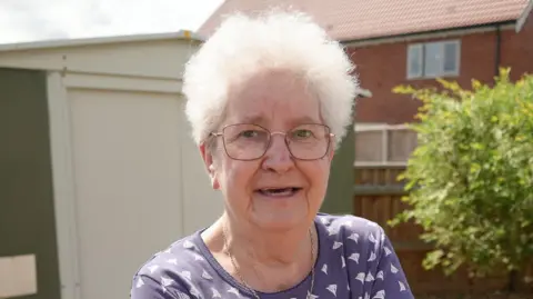 Shaun Whitmore/BBC A head and shoulders image of a woman with short white hair, glasses and a blue top. She is standing in a back garden, with a fence, tree and house behind her. 