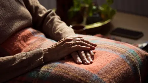 An older person is sitting in a chair with one hand placed on top of another. They have a tartan blanket on their lap.