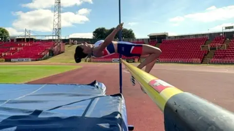 BBC Emily MacDonald high-jumping over a bar, her body parallel to the ground as she clears, in the middle of an athletics stadium surrounded by rows of empty, red seats. A blue landing mat underneath the bar awaits her descent.