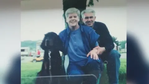 Norrie McIntosh An old photograph of a grey-haired man and woman, crouched down next to a black dog. 