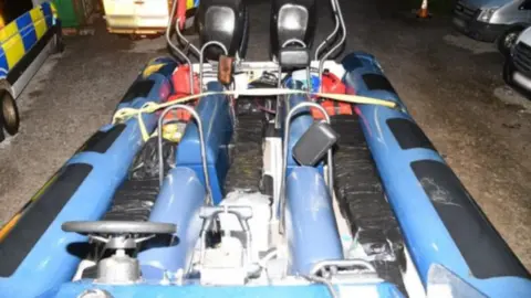 National Crime Agency A blue speedboat that in a car park with several police cars. The boat appears to have a dozen black packages between the seats.