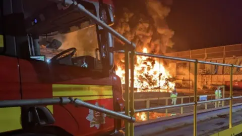 The front of a red fire engine in the foreground, with a large fire and smoke in the background. It is almost white with heat. Two fire fighters seen from behind with helmets and protective gear on. There is a big industrial building or warehouse next to the fire, which is in the open.