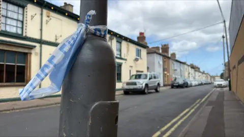 A close-up of part of a torn police cordon tied around a black lamppost. I the background are houses and cars parked at the side of a road.