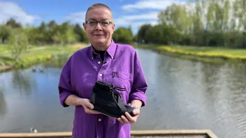 Jeanette Russell smiles and she stands in front of a body of water. It's a sunny day and there is lush green grass on either side with a row of trees and hedges on either side. She has cropped hair and is wearing glasses and a bright purple shirt. She is holding the black lace-up shoe