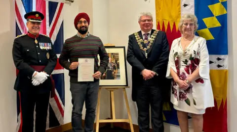 Four people, three men and a woman, standing next to British flags and portraits smiling into the camera. The picture features a man in uniform and the Lord Mayor of Birmingham in his mayoral chains.