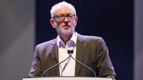EPA/Shutterstock Jeremy Corbyn, who is dressed in a grey suit jacket and white shirt, speaks at a podium with two microphones.