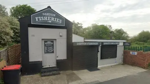 An image of a small building painted black and grey.  There is a small roofed single storey structure to the left and a flat-roofed singled story building attached on the right-hand side.  There are two black roll metal shutters covering windows and the sign stating "Darton Fisheries"