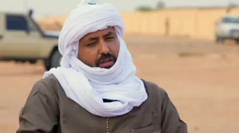 A man sits in front of a car in a sandy opening, wearing a white headscarf.