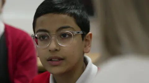 A young schoolboy wearing glasses and a red sweatshirt and polo shirt looks from the camera in a classroom 