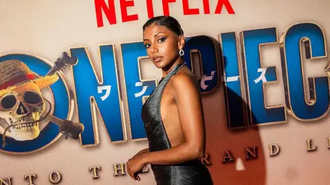 Charithra Chandran wears a metallic strapless dress and has black hair tied up in a bun, with a silver earring. She stands in front of a sign on the red carpet for the One Piece premiere 