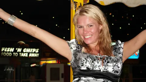 Family of Kirsty Wilkinson Kirsty Wilkinson wears a silver short sleeved dress with a pattern of black flowers on it. She is white, with bleached blonde hair which comes below her shoulders. She has her arms raised and is smiling at the camera. She wears a wide silver bangle. In the background, which is at night, a sign reading "Fish'n'Chips food to take away" is illuminated. There is a yellow pole behind her and the edge of a canopy. It looks like a fairground site.