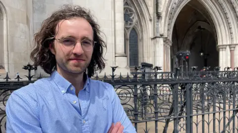 David Hamon has shoulder-length brown hair, is wearing glasses and a light blue shirt with the collar unbuttoned and has his arms crossed as he stands in front of the Royal Courts of Justice in London 