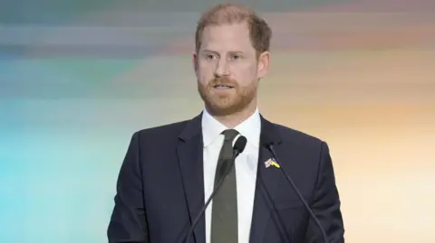 Prince Harry - a man in a dark business suit, white shirt and olive green tie, stands in front of two small microphones as he delivers a speech. The background goes from blue to sunrise yellow in a gentle gradient.