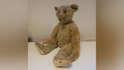 A light brown teddy bear, showing clear signs of use and missing fur. It is sitting upright on a white table. 