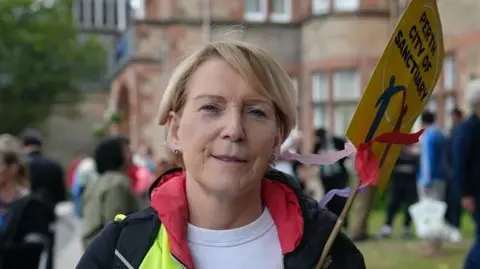 Mary Alexander has short blonde hair and holds a sign which says "Perth City of Sanctuary" outside a hotel with people in the background. She wears a black, yellow and red jacket and a white top.