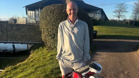 Stewart Owen A man stands with his hands in his pocket next to some golf clubs and with a building and greenery in the background