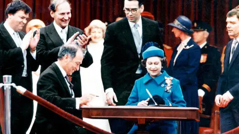 Getty Images The Queen, in a blue suit and hat, sits at a table laid with a pen and a document, while Pierre Elliott Trudeau sits at the other end in a black suit, smiling. They are surrounded by happy looking politicians in suits