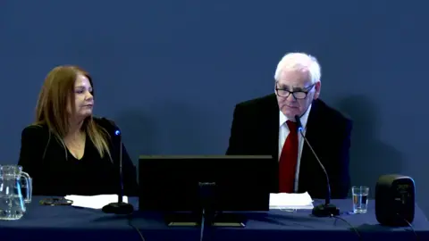 Michael Gallagher gives evidence at the Omagh inquiry with his daughter Cat. Mr Gallagher has grey hair and black rimmed glasses and is wearing a dark suit with a red tie and white shirt. His daughter has long brown hair and is wearing a  dark suit and is looking at her father