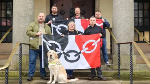 A group of six people are standing on stone steps leading up to large wooden doors. There are handrails either side. Three men are standing in the back row holding a large black, white and red flag with a chain link design on it. There are two men and one woman, who is standing in the middle, in the row in front. They are also holding a flag with the same details on it. There is a large guide dog in front of them. The people in the image are: Karl Denning, Steve Edwards, Karen Westwood, Peter Drake, Pete Lowe, Matt Cook.