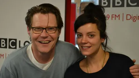 Robert Elms wearing a grey jumper over a white T-shirt with his arm around Lily Allen who is wearing a black top