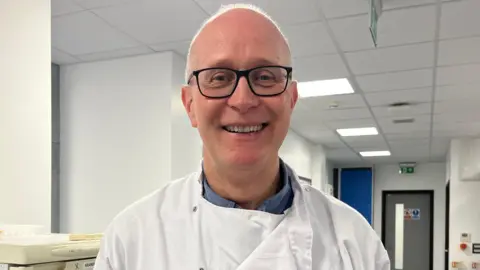 A man in black glasses and a white lab coat smiles into the camera. He is bald and is standing in a laboratory. 