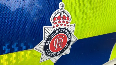 A photo of a Gloucestershire Constabulary on the side of a police care. It is surrounded by blue and yellow reflective stickers