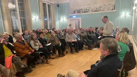 One of the farmers speaking at the public meeting. He is standing besides the other panellists. People in the audience are seen taking notes, pictures and some are drinking water.