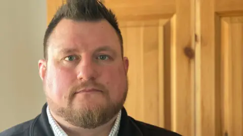 Snagger Chris Greenwood is wearing a checked shirt with a black fleece gilet over the top. The background is a wooden door and pale coloured painted wall.
