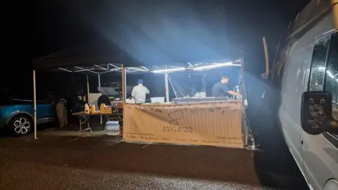 National Grid A welfare van offering hot food and drinks to customers. There are two people behind the catering stand and a white van on the right.