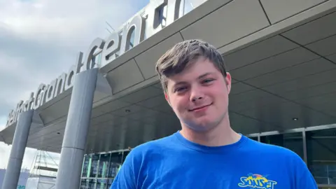 Luke is standing outside the new station. He was brown hair swept to the right side. He's wearing a blue top and smiling at the camera. The station has white pillars holding it up and white lettering saying Grand Central Station. The sky is blue and there are some clouds. 