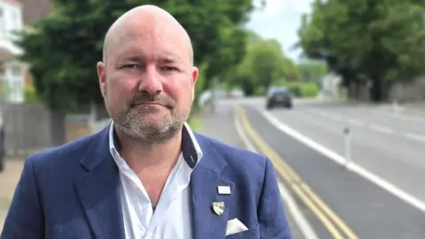 A man in a blue blazer stands on a suburban roadside, facing the camera with hands in pockets. To the left, a flowering shrub borders a low brick wall and houses. To the right, a street with a bike lane, passing cars, and shops stretches into the distance beneath leafy trees and overcast light.