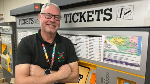 Nexus Alex Carr, with grey hair and glasses, wearing a black T-shirt with a Nexus logo on, has his arms folded across his chest and is leaning on a ticket machine in a station. 