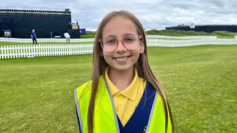 A young girl wears a yellow Hi-Viz vest. Underneath she wears a blue jumper and yellow polo shirt. Behind her is the back of a large navy stand. There is a green grass putting green over her right shoulder. 