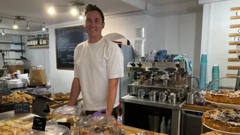 Harry Picken standing in his business. He is standing behind a table with baked goods on it. Behind him there is a coffee machine and a menu board.