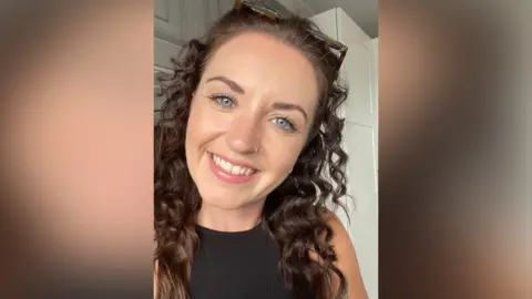 Faye Ramsey A selfie-style image of Faye Ramsey who is smiling at the camera. She has long brown hair which is curly and also has a nose ring on her right hand nostril. Her glasses rest on the top of her head and she is wearing a black sleeveless top.