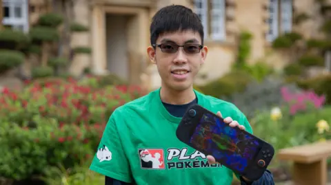 University of Cambridge Marcus Koh is looking at the camera photographed in a garden with plants and flowers blurred behind him. He is holding a gaming device and wearing a green top. He has short, dark hair and is wearing spectacles.