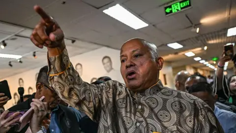 Philippine Senator Ronald Bato Dela Rosa gestures with his right hand, pointing upwards with his index finger while surrounded by journalists in the Senate hall