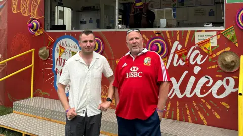 A man in a white, short-sleeved shirt stands next to man in red football T-shirt with sunglasses on his head. They are in front of a large, red food truck with sombrero hats, Mexican-themed bunting and a colourful yellow design on it.