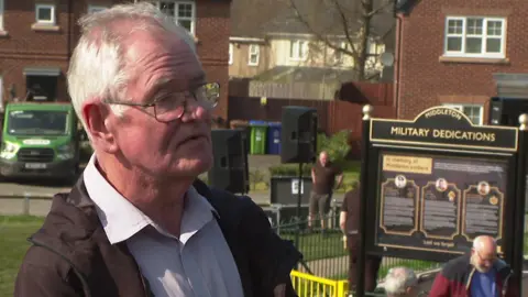 A man with short grey hair, a light coloured shirt and a dark jacket is being interviewed as he stands in front of the memorial sign.
