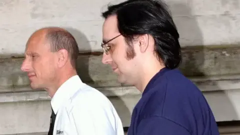 Media Wales Jeffrey Gafoor being led into court by a prison guard. He has swept back dark hair, large sideburns and glasses and is wearing a dark blue jumper