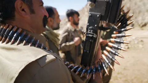 Matthew Goddard/BBC Three Iranian Kurdish fighters stand looking to the right. The nearest one holds an automatic weapon and has an ammunition belt around his neck.