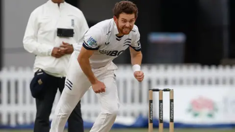 Middlesex's Ryan Higgins, in whites, completes his bowling action with stumps and umpire visible