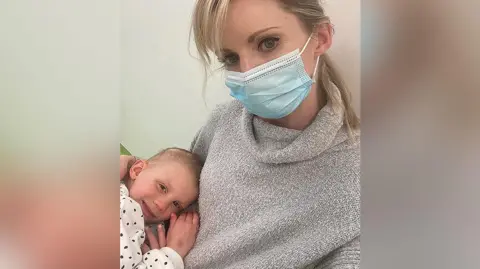 Chloe (left) when she was three cuddling into her mother Nikki. She is wearing a white and black polka dot top. She is smiling. Nikki has blond hair tied back wearing a face mask and beige woolly jumper. 