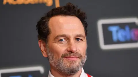 Getty Images Rhys at a press junket wearing a white sweater with red and blue stripes and a white shirt with a red tie 