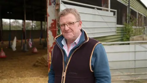 Robert Garner smiles at the camera while standing outside a turkey pen on his farm. He has short light brown hair, glasses and wears a brown gilet jacket with a blue jumper underneath and a shirt. 
