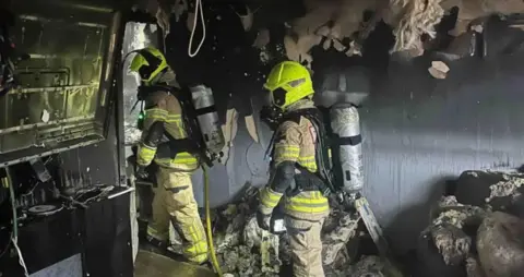 South Wales Fire and Rescue Service Two firefighters in full uniform and wearing yellow helmets and oxygen tanks on their banks in a room gutted by fire. The walls are black and peeling, while a sofa has been badly damaged and there is debris on the floor.